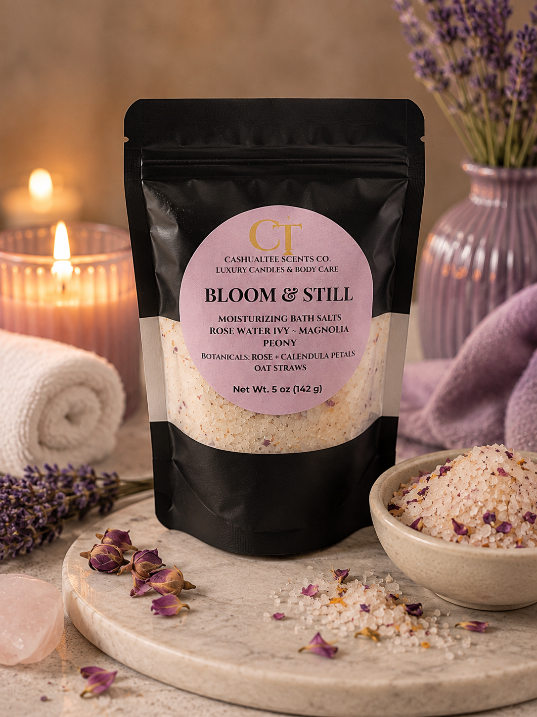A black stand-up pouch labeled 'Bloom & Still' by Cushion Butter Scents Co., surrounded by bath salts, dried lavender, a lit candle, and a rolled towel in a spa-style setting.