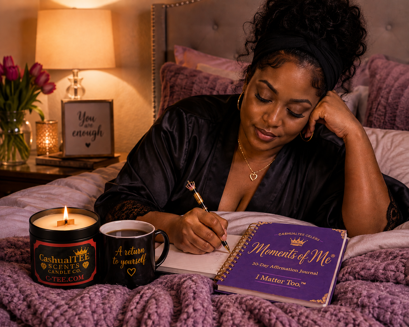 A woman in a black satin robe lies in bed writing in a purple journal titled 'Moments of Me: 50+ One Hour Affirmation Journal I Matter Too™'. A lit candle jar labeled 'Cashmere Soft Candle Co' and a black mug sit beside the journal on a bed with pink bedding and a bedside lamp glowing in the background.