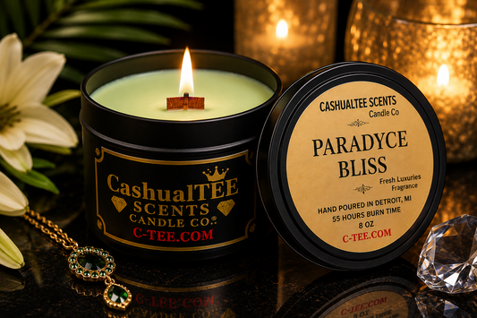 A lit black metal candle tin labeled CashualTEE SCENTS Candle Co. and Paradyce Bliss, shown with its lid beside it on a reflective dark surface, with warm blurred candles and decorative flowers in the background.